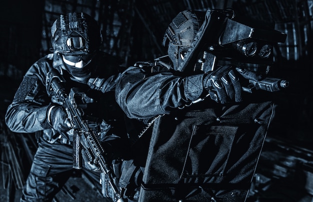 Foto combatientes de grupo táctico de operaciones especiales de la policía con uniforme negro y máscara, pistola armada y rifle de servicio, protegiéndose con un escudo balístico blindado, escondiéndose en la oscuridad durante una redada nocturna