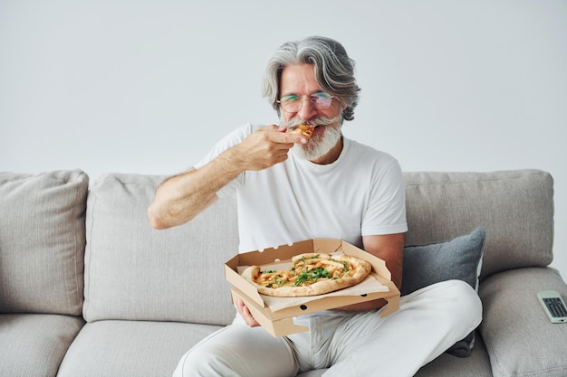 Come deliciosa pizza mientras ve el programa de televisión Senior hombre moderno y elegante con pelo gris y barba en el interior