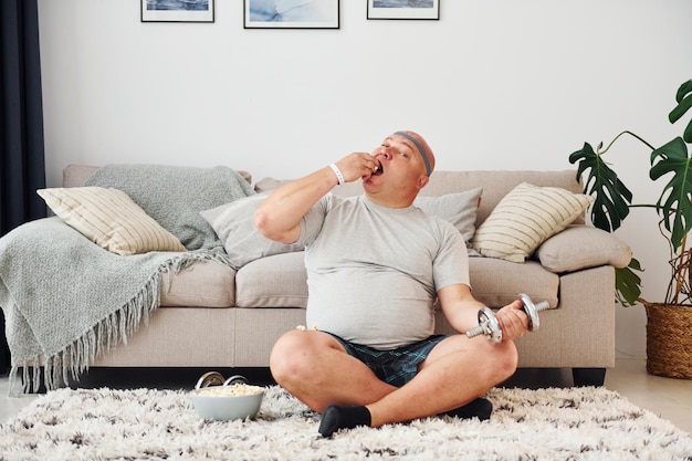 Come e faz exercícios Homem engraçado com excesso de peso em roupas casuais está dentro de casa em casa