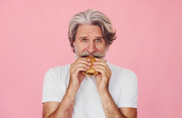 Come um delicioso hambúrguer. Um homem idoso moderno e elegante, com cabelos grisalhos e barba, está dentro de casa.