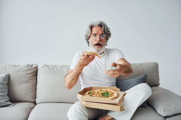 Come uma deliciosa pizza enquanto assiste ao programa de tv homem moderno elegante sênior com cabelos grisalhos e barba dentro de casa