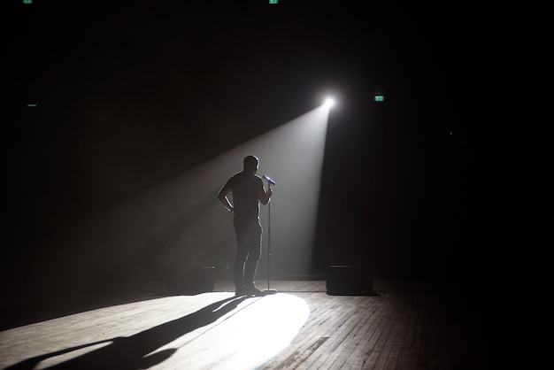 Foto comediante de pé no palco sob o feixe de luz.