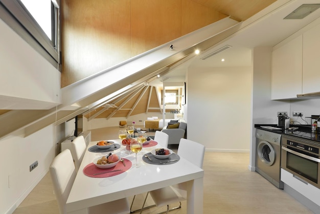 Comedor con mesa de madera blanca sillas tapizadas en polipiel blanca junto a la cocina abierta en apartamento con techo abovedado y tragaluces