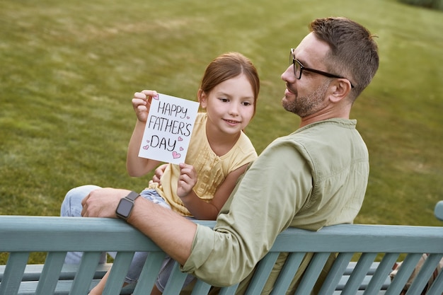 Comemorando o feliz dia dos pais ao ar livre, jovem pai amoroso sentado no banco de madeira no parque com