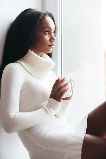 Comenzando el nuevo día con café recién hecho. Mujer africana joven pensativa en suéter caliente mirando a través de una ventana mientras está sentado en el alféizar de la ventana y sosteniendo una taza con bebida caliente