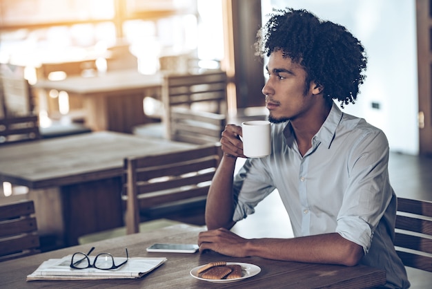 Comenzando un nuevo día en la cafetería. Vista lateral del joven africano sosteniendo la taza de café y mirando a otro lado