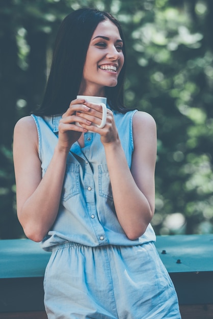 Comenzando su día con una taza de café recién hecho. Hermosa joven bebiendo café y sonriendo mientras está de pie al aire libre con árboles verdes en el fondo