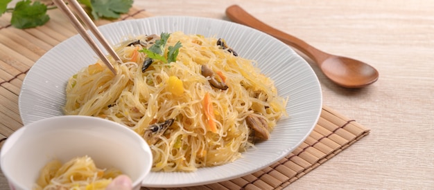 Comer fideos de arroz salteados con calabaza hervida y verduras sobre una superficie de madera.