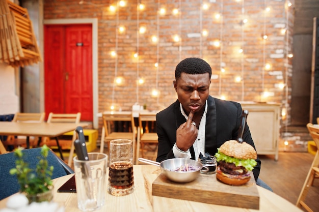 Comer o no Respetable joven afroamericano en traje negro sentado en el restaurante con sabrosa hamburguesa doble y bebida gaseosa