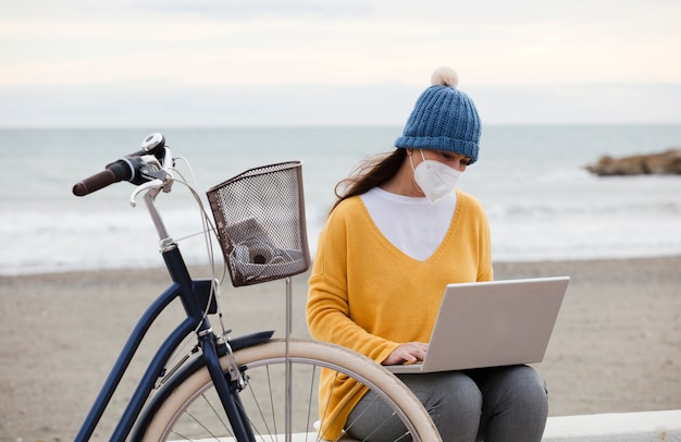 Un comercializador digital que usa una máscara facial está trabajando en un nuevo proyecto de promoción de computadoras portátiles mientras está sentado en el paseo marítimo con una bicicleta. la mujer independiente se comunica con los clientes.