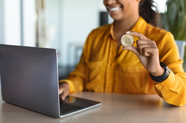 Comercio de criptomonedas Empresaria negra irreconocible sentada en el escritorio con una computadora portátil y mostrando bitcoin dorado