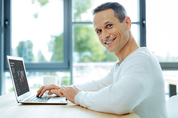 Comercio global. Hombre positivo inteligente alegre usando una computadora portátil y trabajando mientras estudia el comercio global