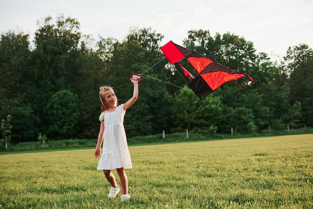 Foto cometa de color rojo y negro. niña feliz vestida de blanco se divierte en el campo. hermosa naturaleza.