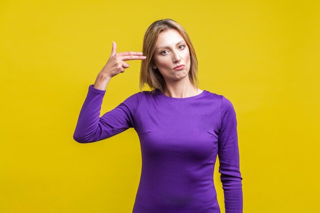 Cometer suicídio Retrato de mulher infeliz no vestido roxo em pé com arma de dedo apontada para a cabeça entediada ou cansada difícil de lidar com o problema tiro de estúdio interno isolado em fundo amarelo