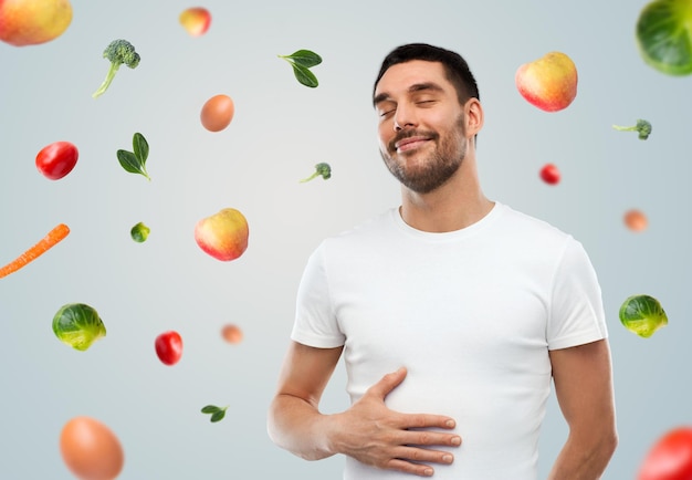 comida, alimentación saludable, dieta y concepto de personas - hombre completo feliz tocando su barriga sobre fondo gris con frutas que caen