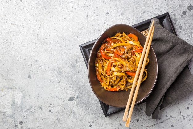 Comida asiática, macarrão udon frito com carne e vegetais em uma tigela em uma superfície de pedra cinza, vista de cima