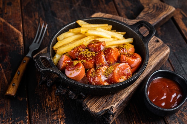 Comida callejera con currywurst, especias de curry en salchichas servidas con papas fritas en una sartén. Fondo de madera oscura. Vista superior.