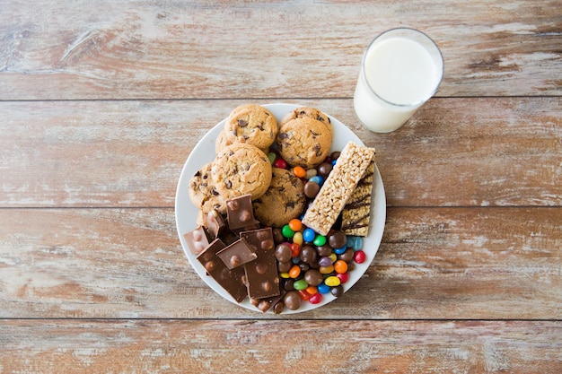 Comida chatarra, dulces y concepto de alimentación poco saludable: cierre de dulces, chocolate, muesli y galletas con vaso de leche en el plato