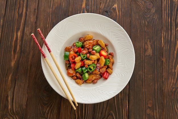 Comida chinesa, frango kung pao em prato branco com pauzinhos na mesa de madeira, vista de cima e espaço de cópia