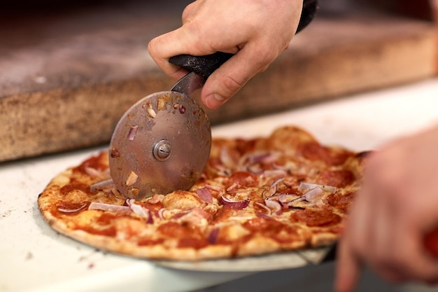 comida, cocina italiana y concepto de cocina: cocine las manos con un cortador cortando pizza en pedazos en la pizzería