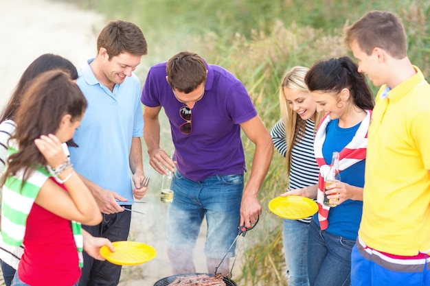 comida, comida, cocina, vacaciones de verano y concepto de personas - grupo de amigos haciendo picnic y haciendo barbacoa en la playa