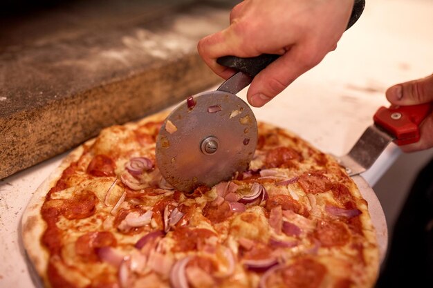 comida, cozinha italiana e conceito de culinária - cozinhe as mãos com cortador cortando pizza em pedaços na pizzaria