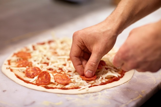 Foto comida, culinaria, cocina italiana, gente y concepto de cocina: manos de cocinero añadiendo rodajas de salami a la pizza en la pizzería