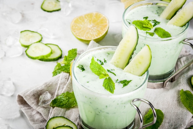 Comida de verão pratos refrescantes sopa fria de pepino abacate com ervas e hortelã com taças com fatias de pepino em uma mesa de concreto branca com ingredientes