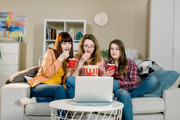 Comida e filmes. Três irmãs bonitas emocionais comendo pipoca ao assistir filme ou programa de esporte enquanto está sentado no sofá da sala bonita em casa.
