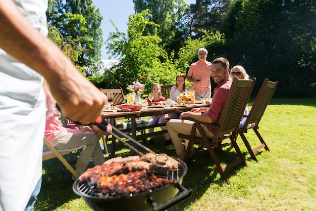 Foto comida, gente y concepto de tiempo familiar - hombre cocinando carne en la parrilla de barbacoa en una fiesta de jardín de verano