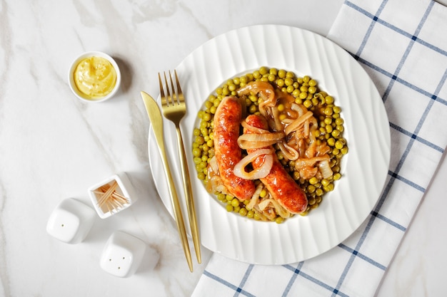 Comida inglesa en un plato blanco: salchicha de cerdo asada con guisantes y salsa de cebolla clásica sobre una mesa de mármol blanco con cubiertos dorados y mostaza inglesa, vista superior, primer plano