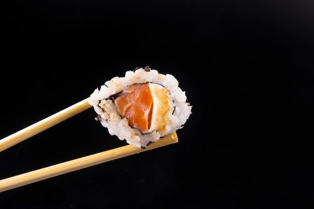 Comida japonesa caseira segurando sushi de salmão com creme de queijo com pauzinhos isolados na paisagem macro de fundo preto com molho de soja