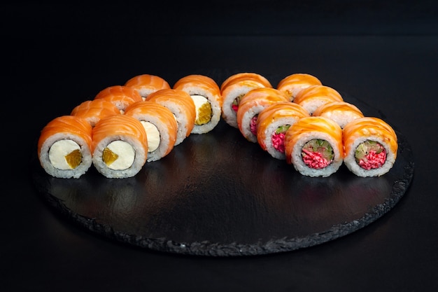 Foto comida japonesa, un juego de rollos con salmón, requesón filadelfia, naranja, pepino, surimi sobre fondo negro.