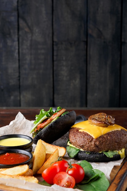 Foto comida para llevar en bandeja de madera en el fondo de madera, de cerca, copie el espacio. hamburguesa de pan negro con tomates, rodajas de papa al horno y salsas para mojar, concepto de comida rápida