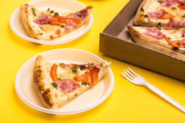 Comida para llevar y entrega. Rebanada de pizza en plato de plástico desechable, caja de pizza y tenedor sobre fondo amarillo