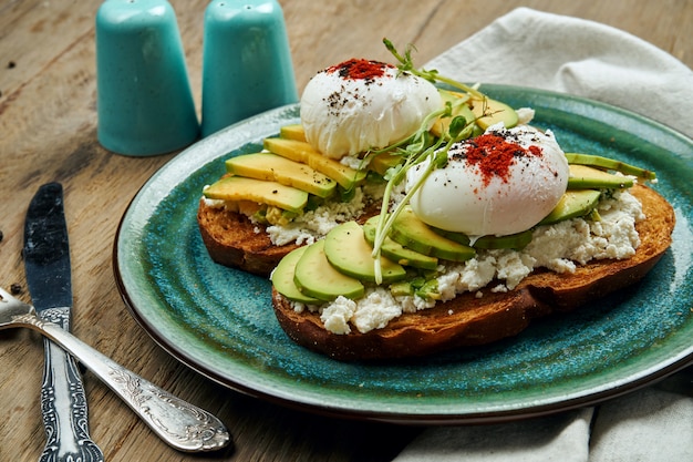 Comida de moda: tostadas de aguacate con ricotta, huevo escalfado sobre pan de centeno en un plato de cerámica sobre una superficie de madera. Comida saludable para el desayuno