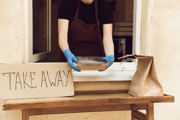 Foto comida. mujer preparando bebidas y comidas, vistiendo guantes y mascarilla protectora. servicio de entrega sin contacto durante la pandemia de coronavirus en cuarentena. quitar concepto. tazas, paquetes reciclables.