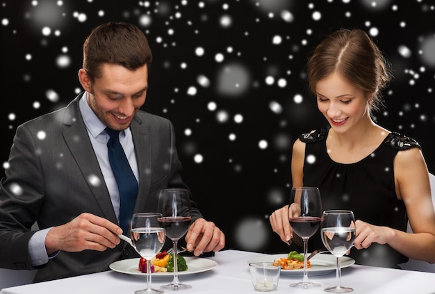 comida, natal, feriados e conceito de pessoas - casal sorridente comendo prato principal no restaurante sobre fundo preto nevado