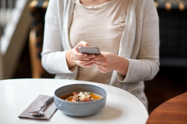 comida, nueva cocina nórdica, tecnología, comida y concepto de personas - mujer sentada en la mesa de café con smartphone y tazón de sopa de calabaza y jengibre vegetal con queso de cabra y ensalada de tomate con yogur