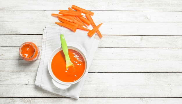 Comida para bebê purê de cenouras frescas com uma colher em um fundo branco de madeira