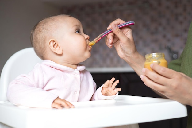 Comida para bebê Um bebê em uma cadeira alta come purê de legumes de uma colher Mãe alimenta o bebê de uma jarra Cozinha Estilo de vida