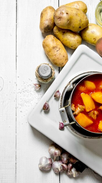 Comida de patata. Patatas guisadas con carne y especias en una cocina eléctrica sobre mesa de madera blanca. Vista superior