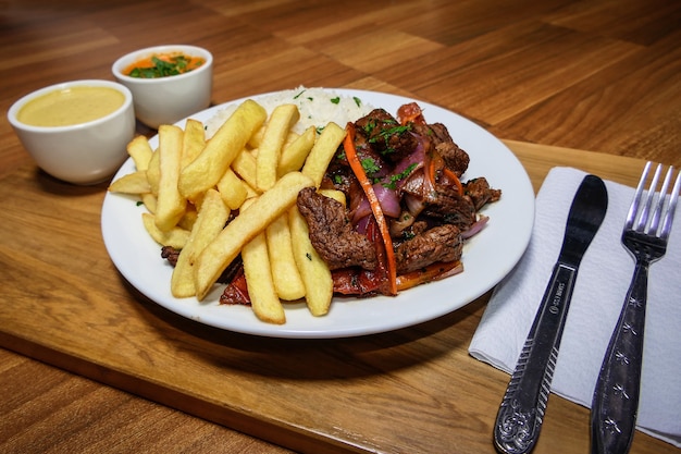 Comida peruana Lomo Saltado, papas fritas con carne, cebolla, pimiento amarillo, perejil, arroz blanco y salsa de soja, sobre un plato blanco con cubiertos y fondo de madera.