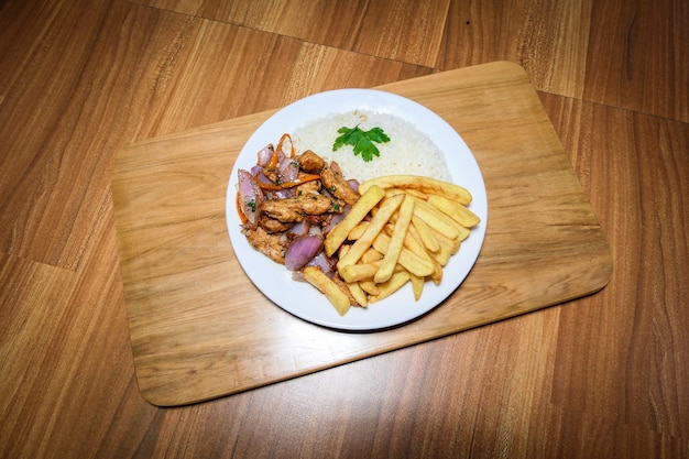 Foto comida peruana pollo saltado, papas fritas, pollo sazonado, cebollas, tomates, verduras asadas, arros blancos en un plato blanco.