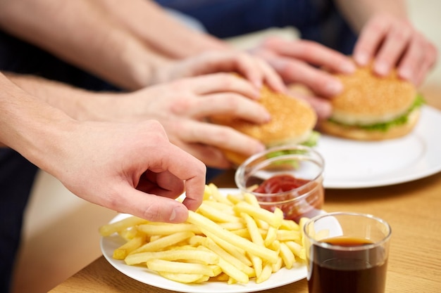 comida rápida, alimentación poco saludable, gente y comida chatarra - cierre de manos masculinas con papas fritas y hamburguesas en la mesa en casa