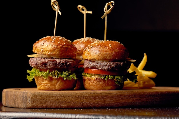 Comida rápida americana tres pequeñas hamburguesas con queso en un tablero. Fondo de madera oscura. Comer con los dedos