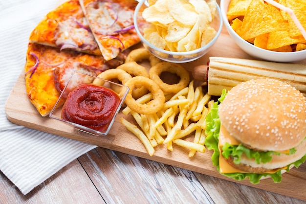 comida rápida y concepto de alimentación poco saludable: cierre de hamburguesa o hamburguesa con queso, anillos de calamar fritos, papas fritas, pizza y ketchup en la vista superior de la mesa de madera