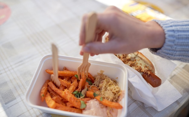 comida rápida, gente y concepto de alimentación poco saludable - cierre de la mano con hot dog y batata en un plato desechable al aire libre