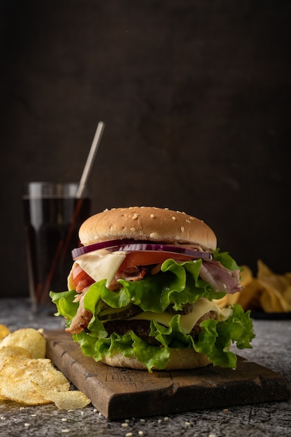 Comida rápida. una hamburguesa grande y jugosa con empanada de ternera, lechuga iceberg, tomates, pepinillos, cebolla y salsa y rebanadas de tocino. Estado de ánimo oscuro y discreto