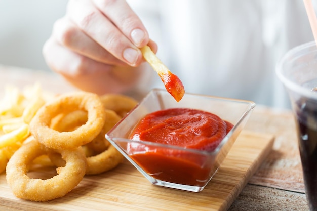 Comida rápida, personas y concepto de alimentación poco saludable: cierre de la mano con anillos de calamar fritos, inmersión de papas fritas en un tazón de ketchup sobre una mesa de madera